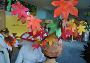 Zbliżenie na narycie głowy jednego z dzieci: czapka wykonana jest z brązowego papieru i wystają z niej ozdobione dużymi kartonowymi liśćmi dwie gałązki.
